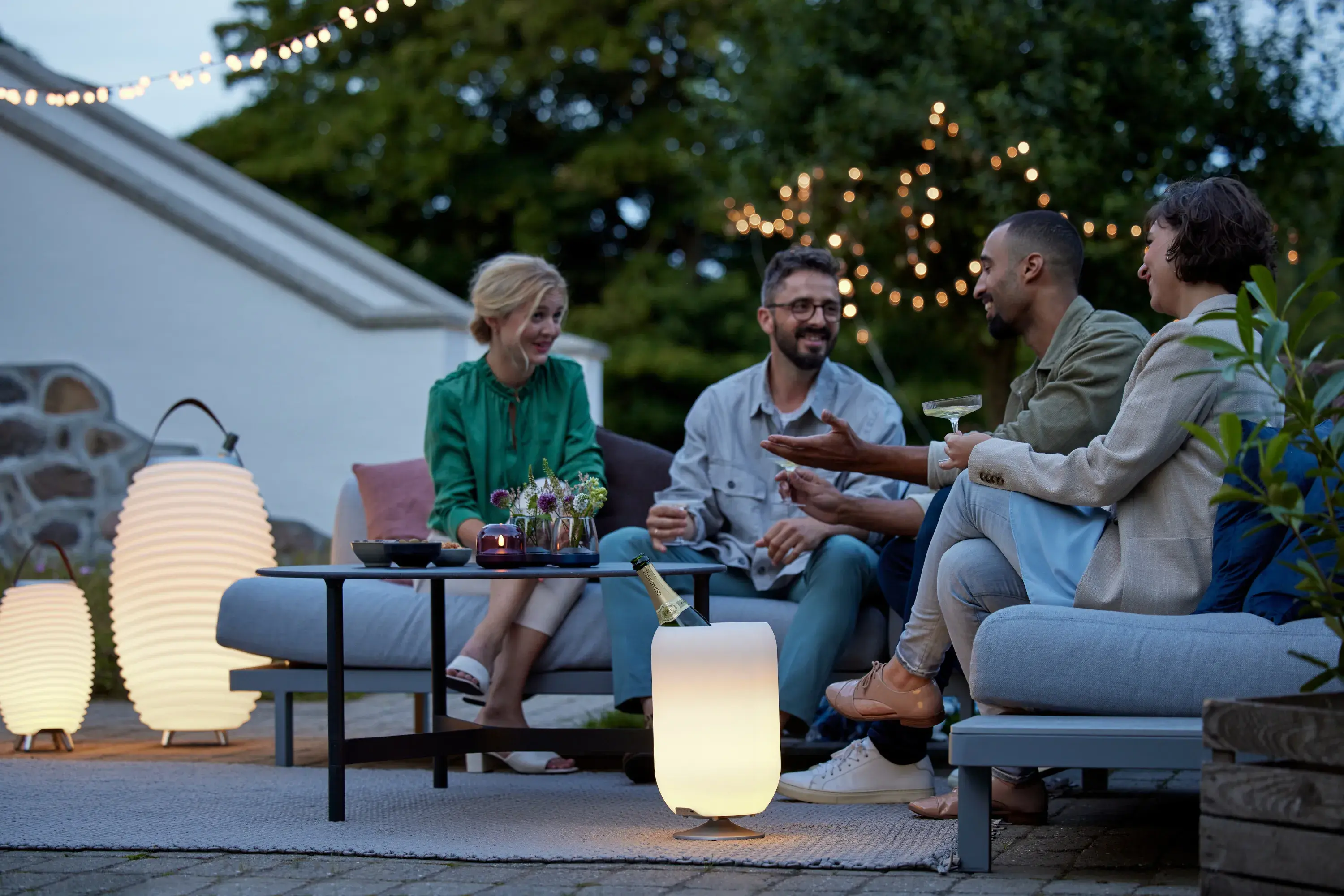 Vier Menschen bei Dämmerung auf gemütlich eingerichteter Terrasse mit verschiedenen Leuchten